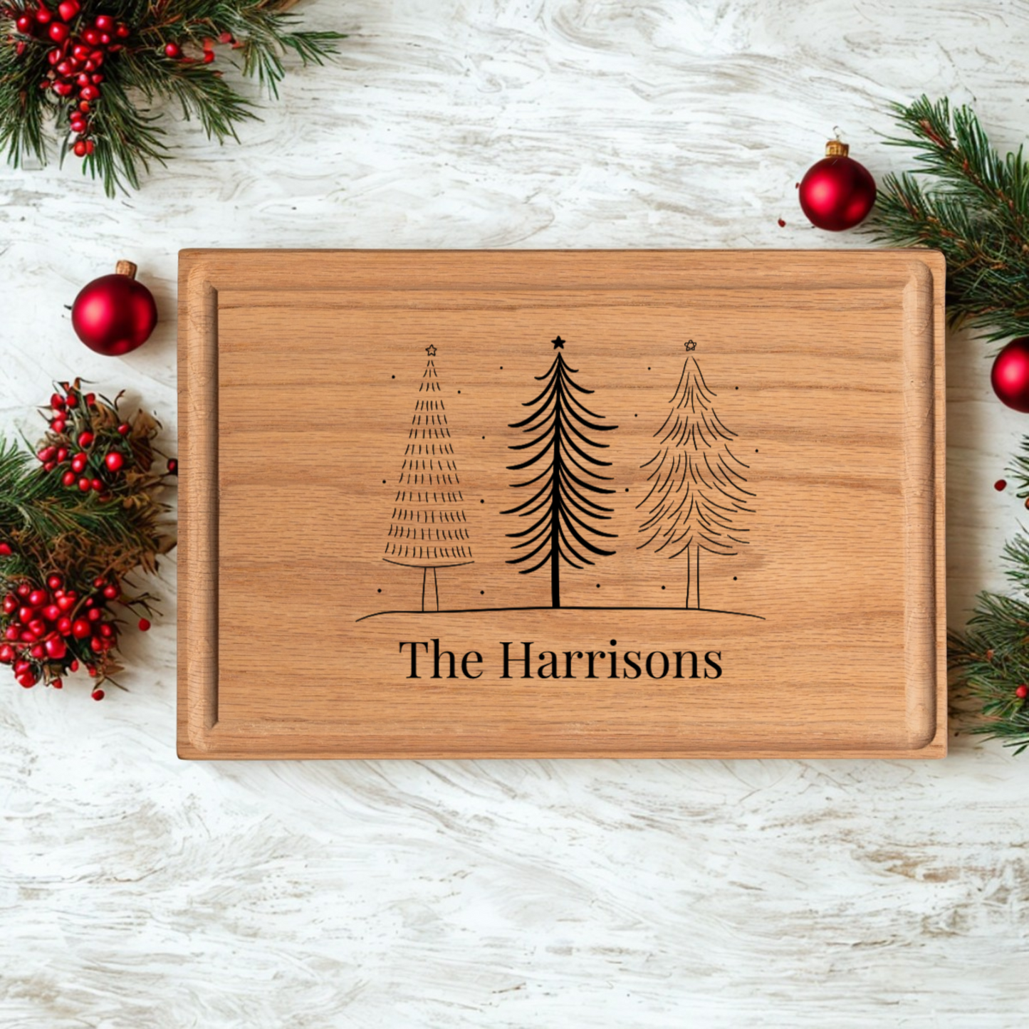 A holiday-themed personalized cutting board crafted from warm oak wood engraved with three stylized Christmas trees and the name “The Harrisons.” The board is placed on a whitewashed wooden surface surrounded by pine branches and red ornaments. This elegant custom cutting board doubles as a holiday serving board and makes a thoughtful cutting board gift for family celebrations.