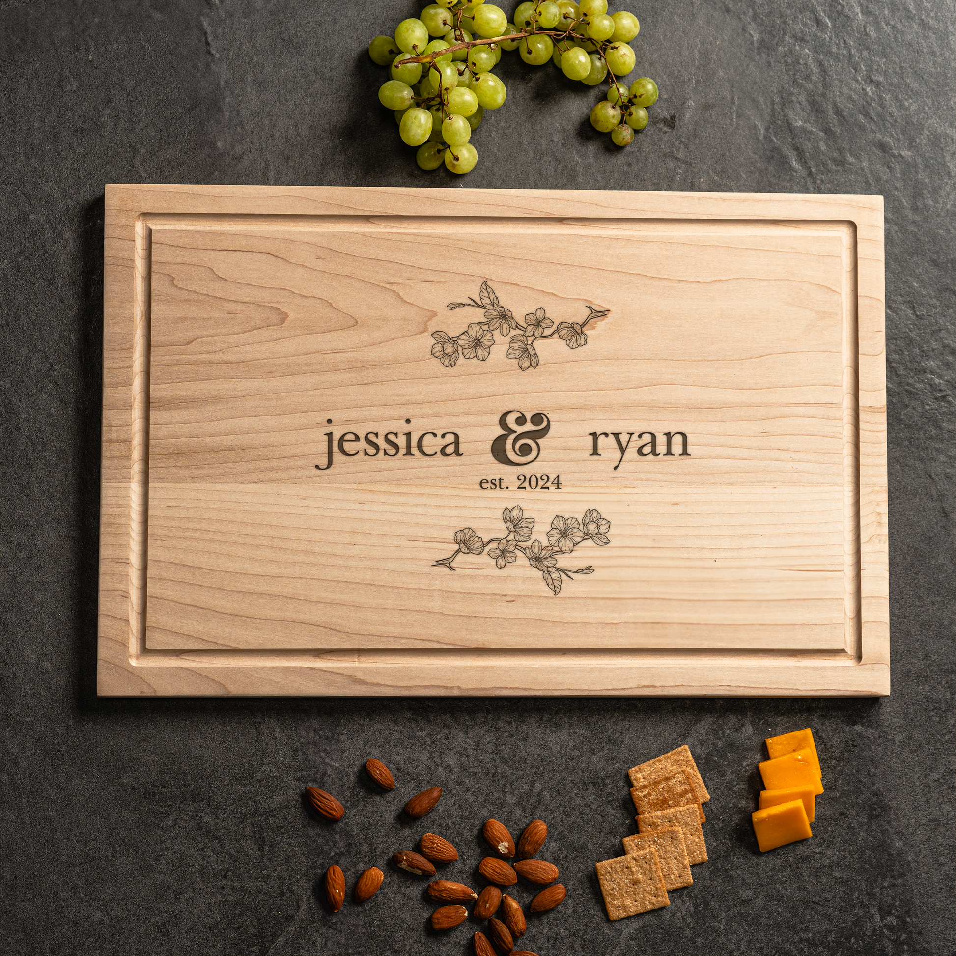 A personalized cutting board made of light maple wood featuring delicate floral engravings and the names “jessica & ryan” with “est. 2024” below. The board is styled on a dark stone surface with green grapes, almonds, crackers, and cheddar cheese cubes nearby. This elegant design highlights the charm of custom cutting boards, making it an ideal wedding, anniversary, or housewarming cutting board gift for couples.