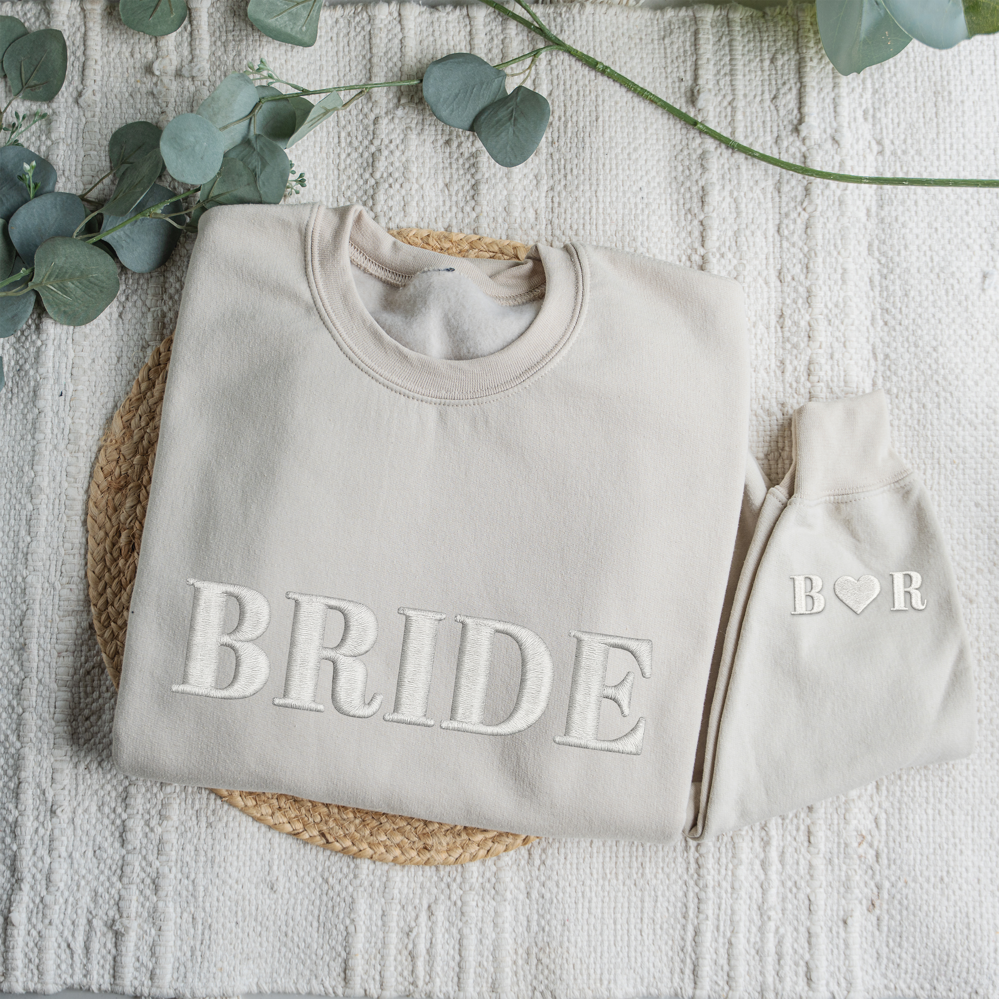 Gray sweatshirt with 'BRIDE' text and matching beanie on a textured surface with eucalyptus leaves.