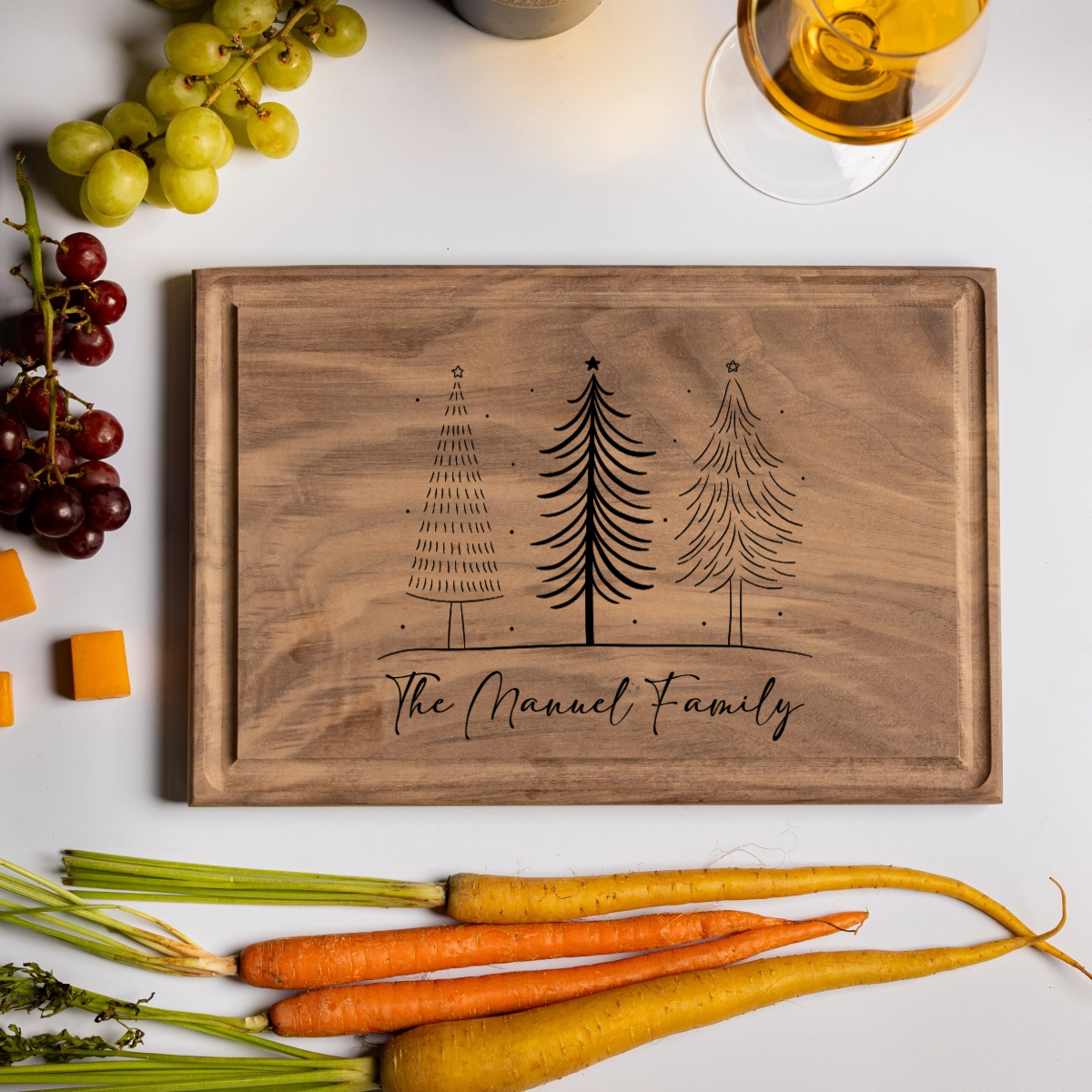 A walnut personalized cutting board engraved with three Christmas trees and the words “The Manuel Family.” Styled with holiday foods—grapes, carrots, cheddar cubes, crackers, and a glass of white wine—this festive board evokes cozy holiday gatherings. A beautiful and practical custom cutting board gift perfect for Christmas entertaining, weddings, or family keepsakes.