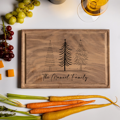 A walnut personalized cutting board engraved with three Christmas trees and the words “The Manuel Family.” Styled with holiday foods—grapes, carrots, cheddar cubes, crackers, and a glass of white wine—this festive board evokes cozy holiday gatherings. A beautiful and practical custom cutting board gift perfect for Christmas entertaining, weddings, or family keepsakes.