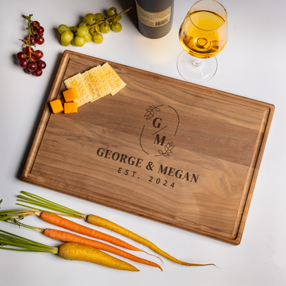 A personalized cutting board crafted from rich walnut wood engraved with the names “George & Megan” and “Est. 2024,” accented by a monogram “G M” surrounded by leafy botanical details. The board rests on a white surface with green and red grapes, a bottle of wine, a glass of golden white wine, crackers, cubes of cheddar cheese, and heirloom carrots. This elegant custom cutting board doubles as a stylish serving board and makes a timeless cutting board gift for weddings, anniversaries, and housewarmings.