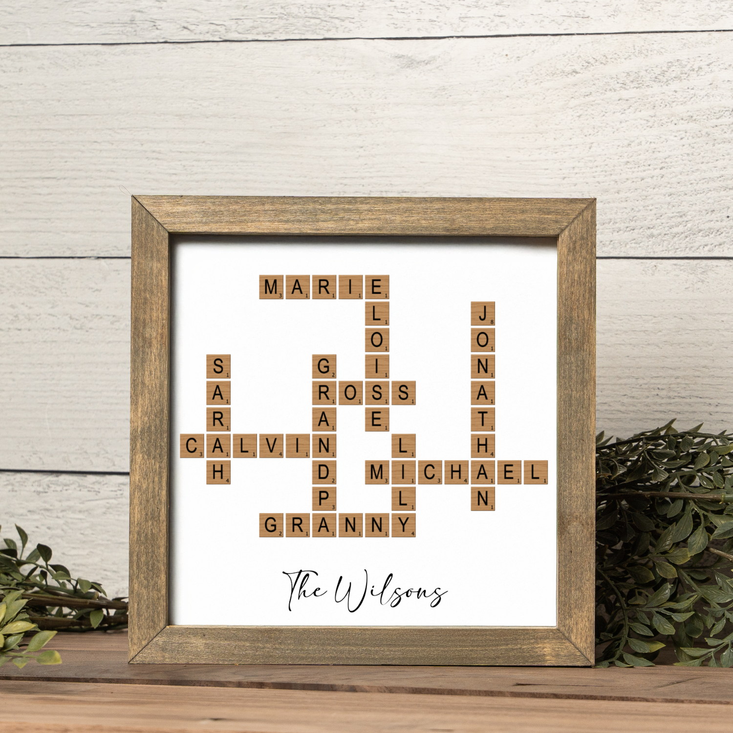 A custom crossword family name frame featuring scrabble-style wooden tiles arranged to spell multiple family names including “MARIE,” “CALVIN,” “JONATHAN,” and “GRANNY.” The text “The Wilsons” is elegantly written in black script beneath the design. Displayed in a rustic walnut-toned frame with greenery accents, this personalized family crossword art makes a unique home décor accent and a heartfelt gift for grandparents, parents, or family celebrations.