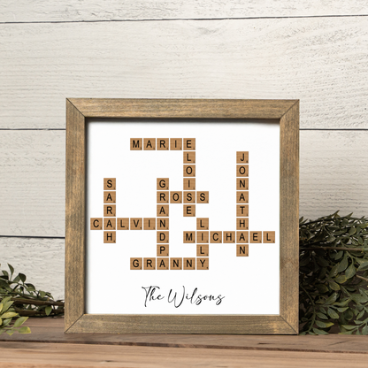 A custom crossword family name frame featuring scrabble-style wooden tiles arranged to spell multiple family names including “MARIE,” “CALVIN,” “JONATHAN,” and “GRANNY.” The text “The Wilsons” is elegantly written in black script beneath the design. Displayed in a rustic walnut-toned frame with greenery accents, this personalized family crossword art makes a unique home décor accent and a heartfelt gift for grandparents, parents, or family celebrations.