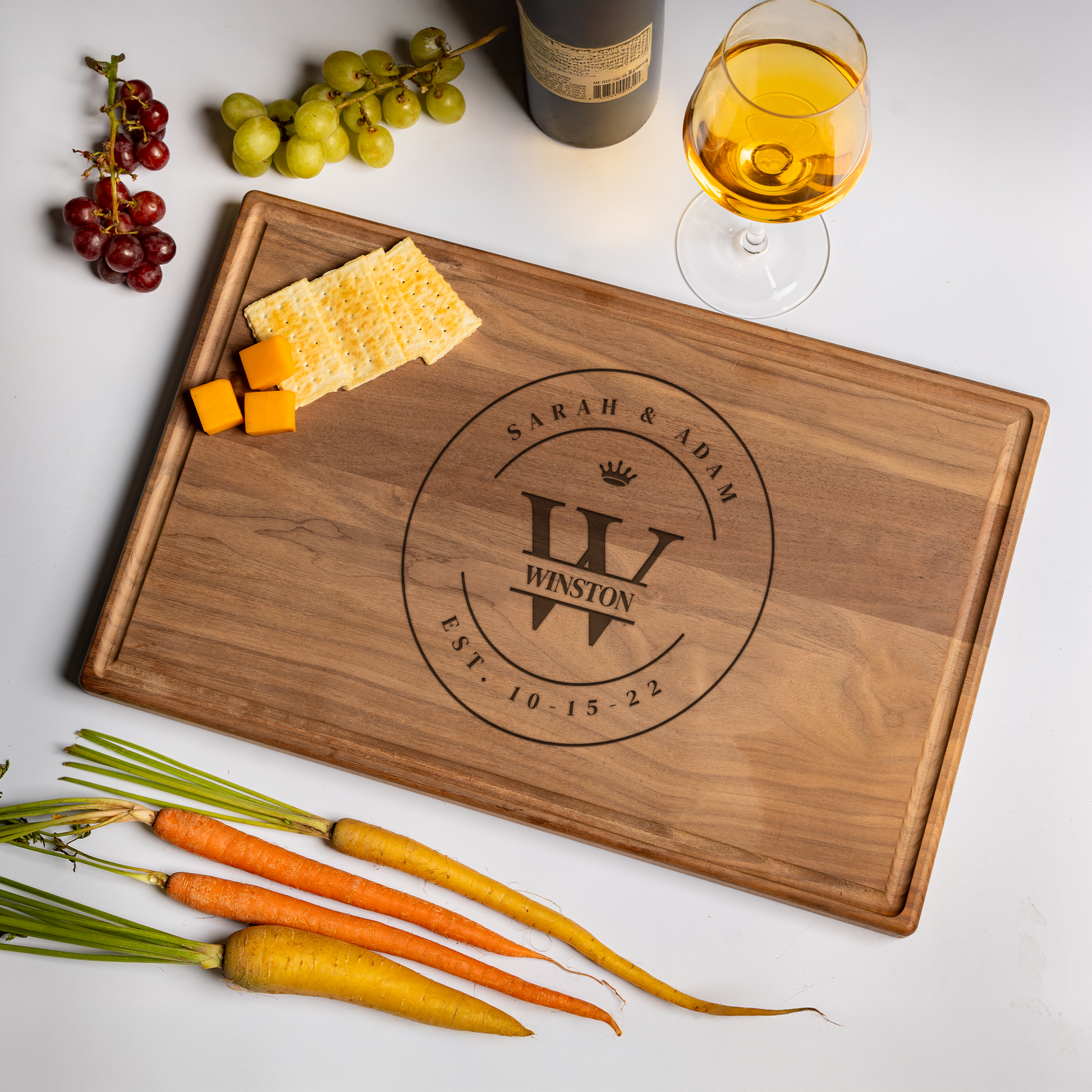 A personalized cutting board made of luxurious walnut wood engraved with a large monogram “W” topped with a crown, the family name “Winston” across the center, and “Sarah & Adam” with “Est. 10.15.22” encircling the design. The board is styled on a clean white background beside red and green grapes, a wine bottle, a glass of golden wine, cheddar cheese cubes, crackers, and fresh heirloom carrots. This elegant custom cutting board makes the perfect cutting board gift for weddings, anniversaries, or homeowners