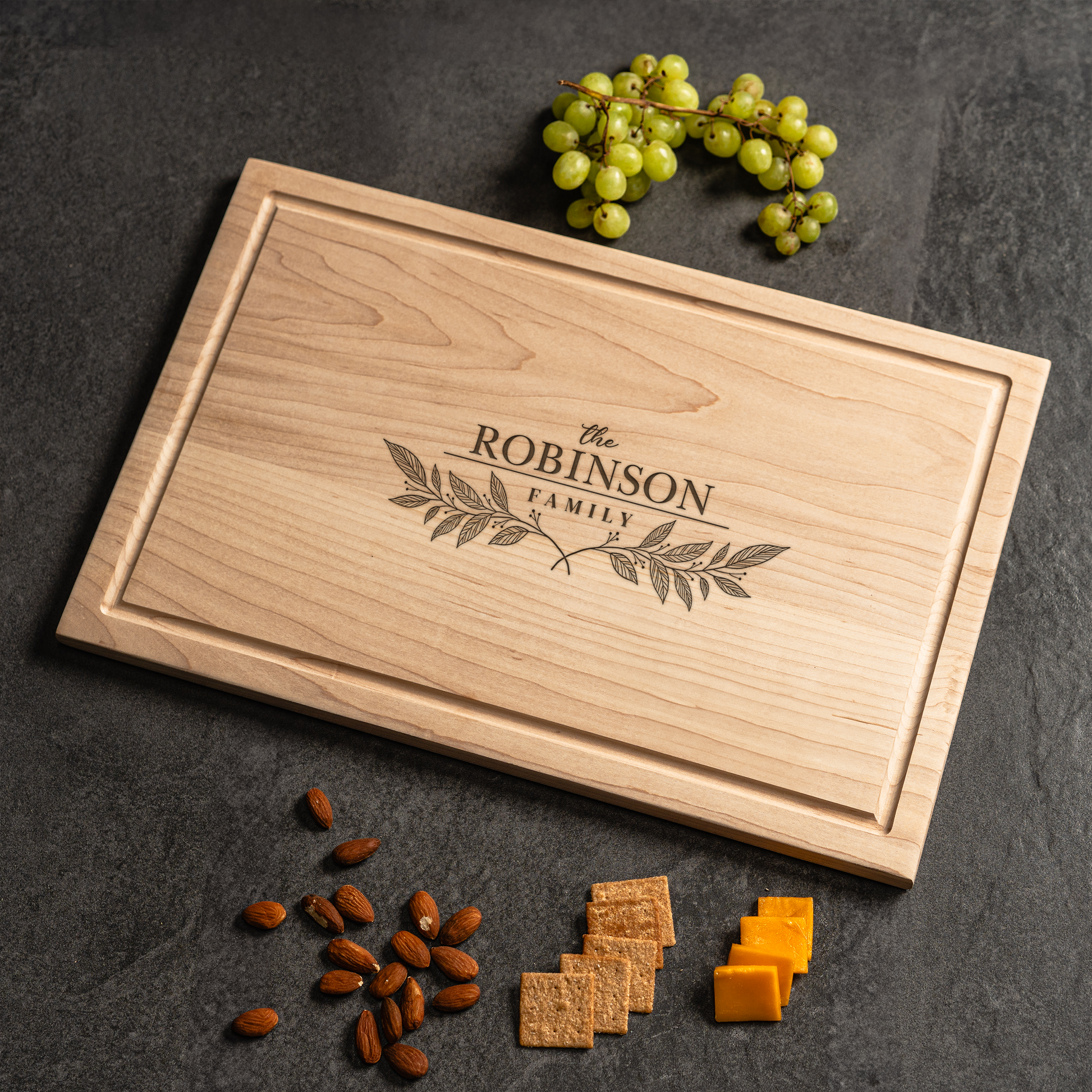 A personalized cutting board made from light maple wood engraved with “The Robinson Family” surrounded by elegant leafy branches. The board is styled on a dark stone surface with a cluster of green grapes, almonds, wheat crackers, and cubes of cheddar cheese arranged nearby. This timeless custom cutting board is perfect for weddings, anniversaries, and family celebrations, serving as both a functional kitchen piece and a meaningful cutting board gift for loved ones.